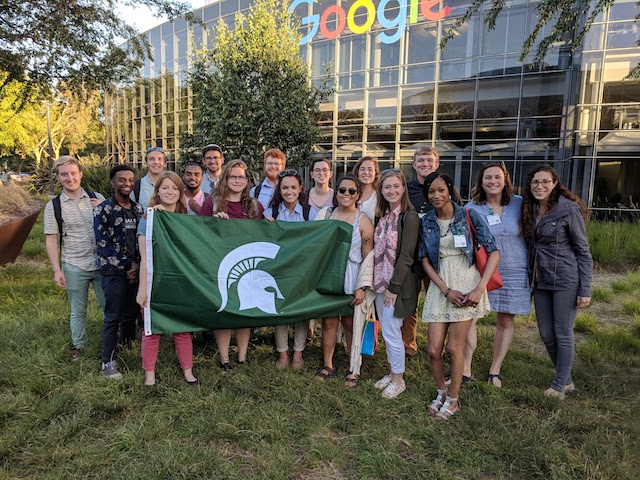 Group standing in front of Google's headquarters, holding a Michigan State flag.