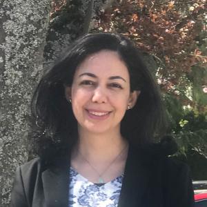 Yasmine has dark brown hair and brown eyes. She has a patterned top with a black cardigan on top. She is smiling at the camera