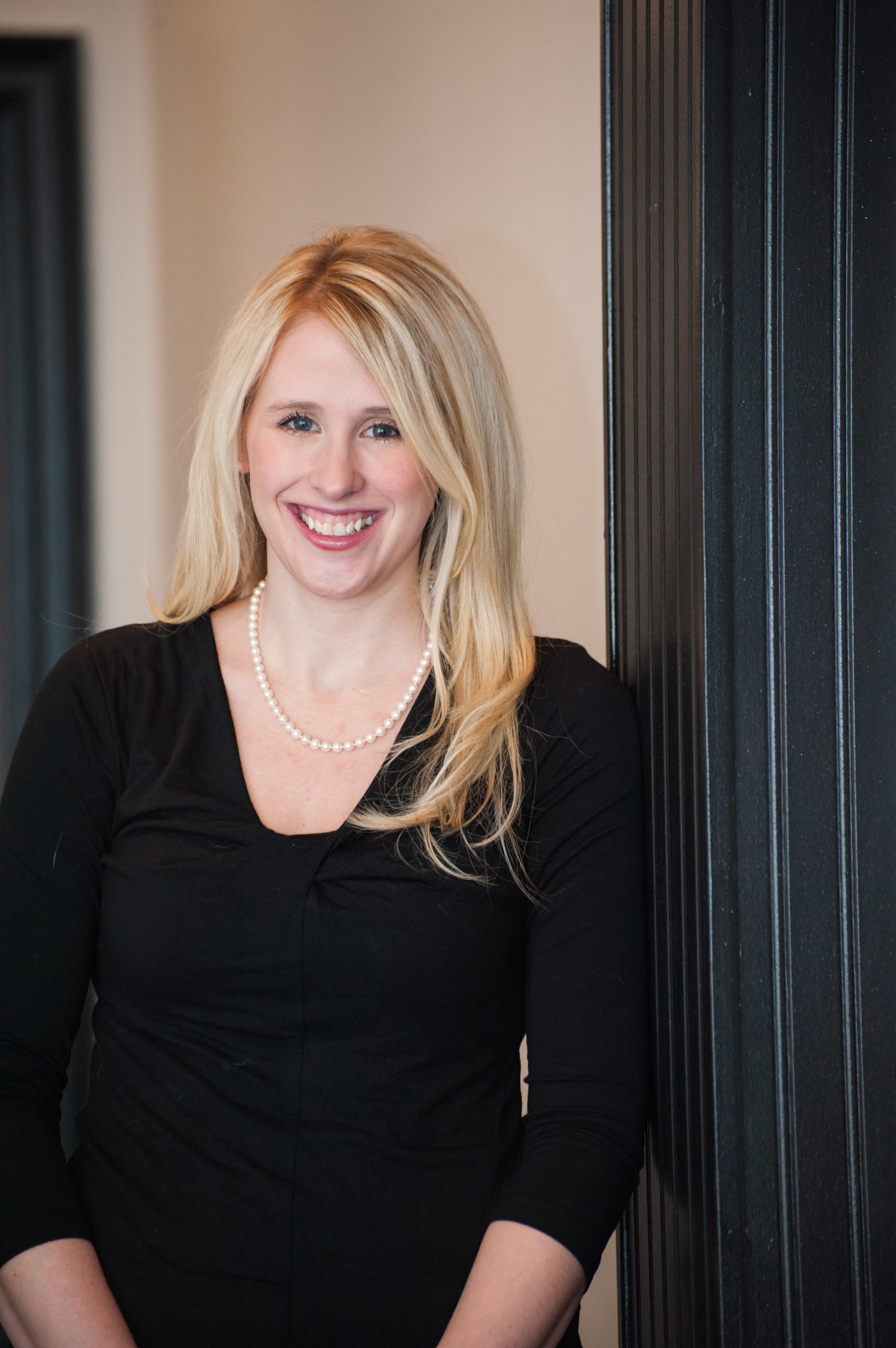 Leslie has long blonde hair and blue eyes. She is wearing a black dress with a pearl necklace and pink lipstick. She is smiling at the camera