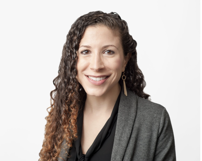 Laura has curly long brown hair and brown eyes. She is wearing a black top with a grey blazer. She is smiling at the camera