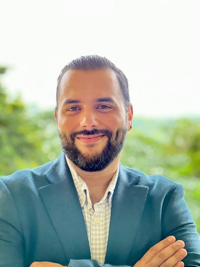 Ronaldo has slicked-back brown hair and a beard. He has dark brown eyes and is smiling for the camera. He is wearing a teal suit coat with a patterned shirt underneath.