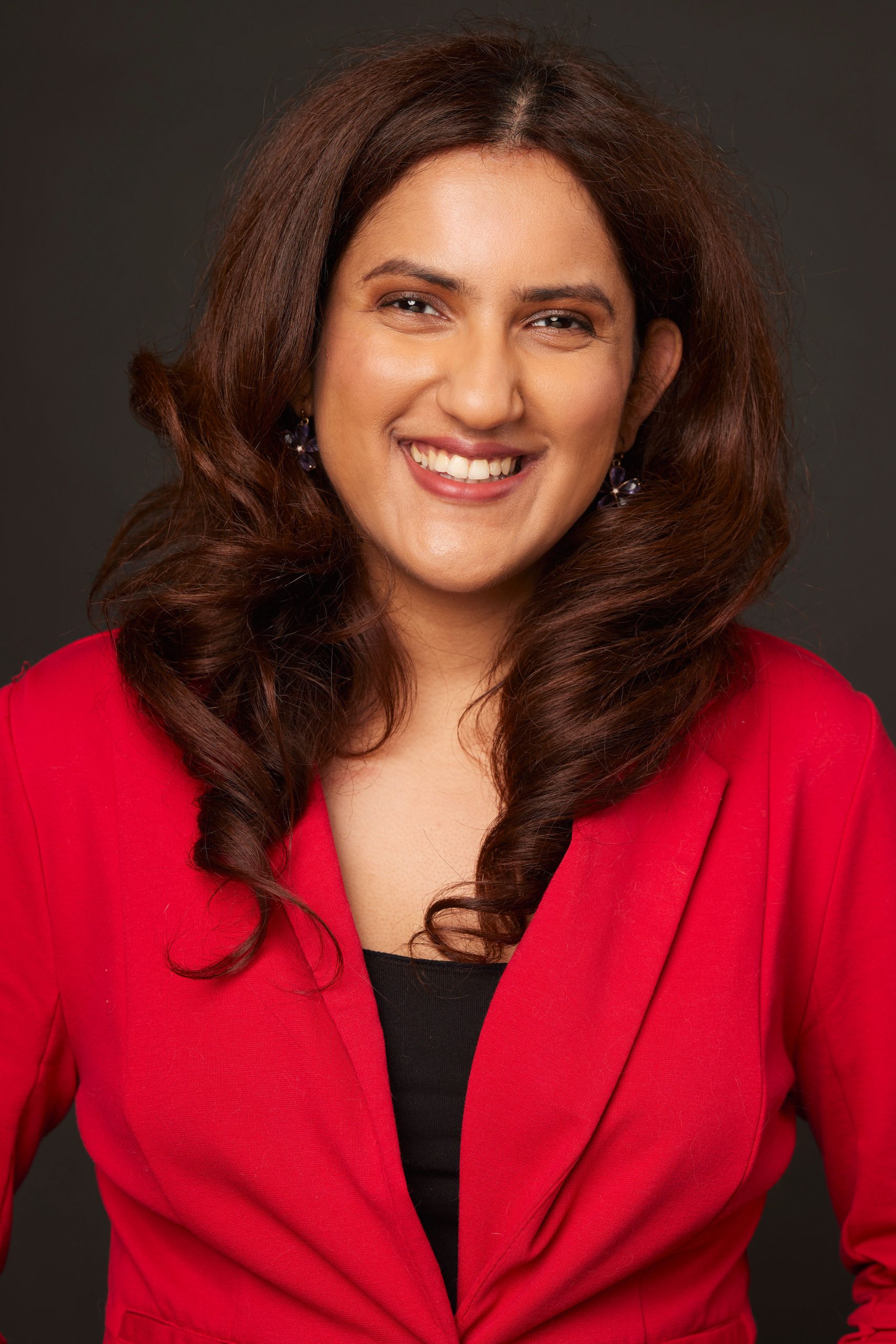 Meena has medium-toned skin and collarbone-length dark brown hair. She is smiling looking directly at the camera and wearing red blouse.