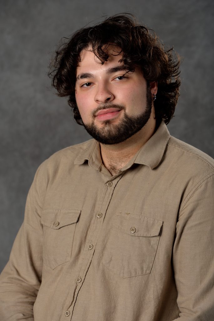 Julius Patto has medium-length dark brown hair, and a short beard. He is smiling at the camera wearing a wheat button-up. There is a grey background behind him.