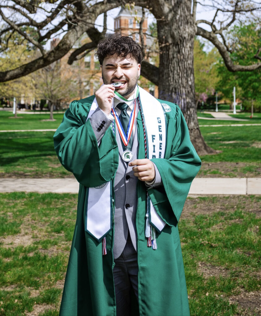 Julius standing outside wearing green graduation gown with first generation sash and medals.