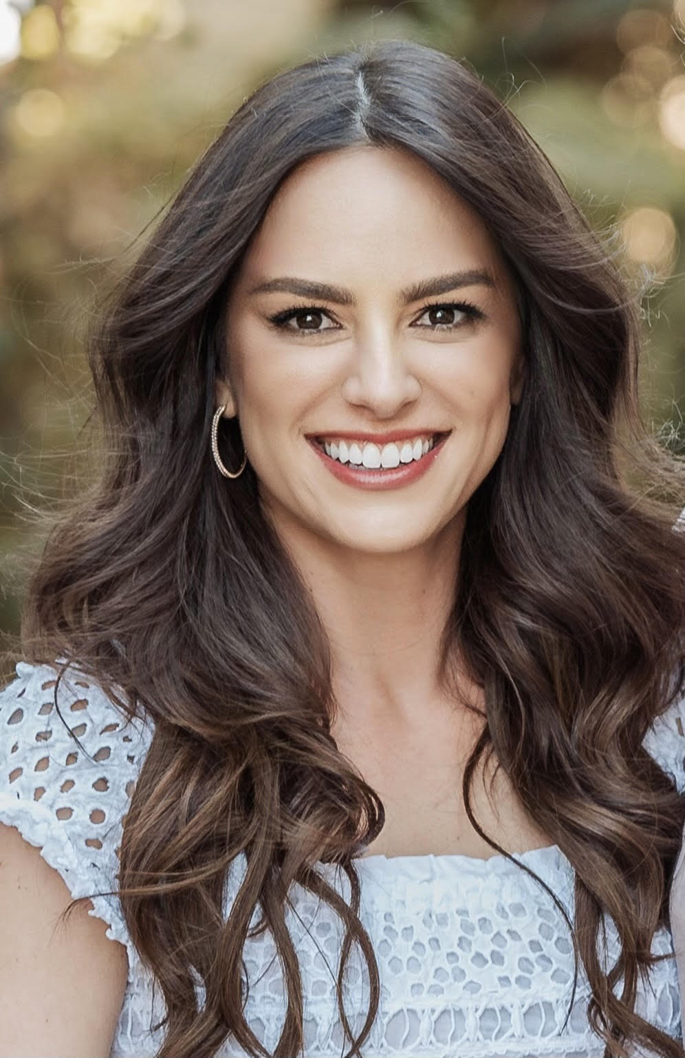 Liz has long dark brown hair, brown eyes, wearing a white top, and smiling at the camera.