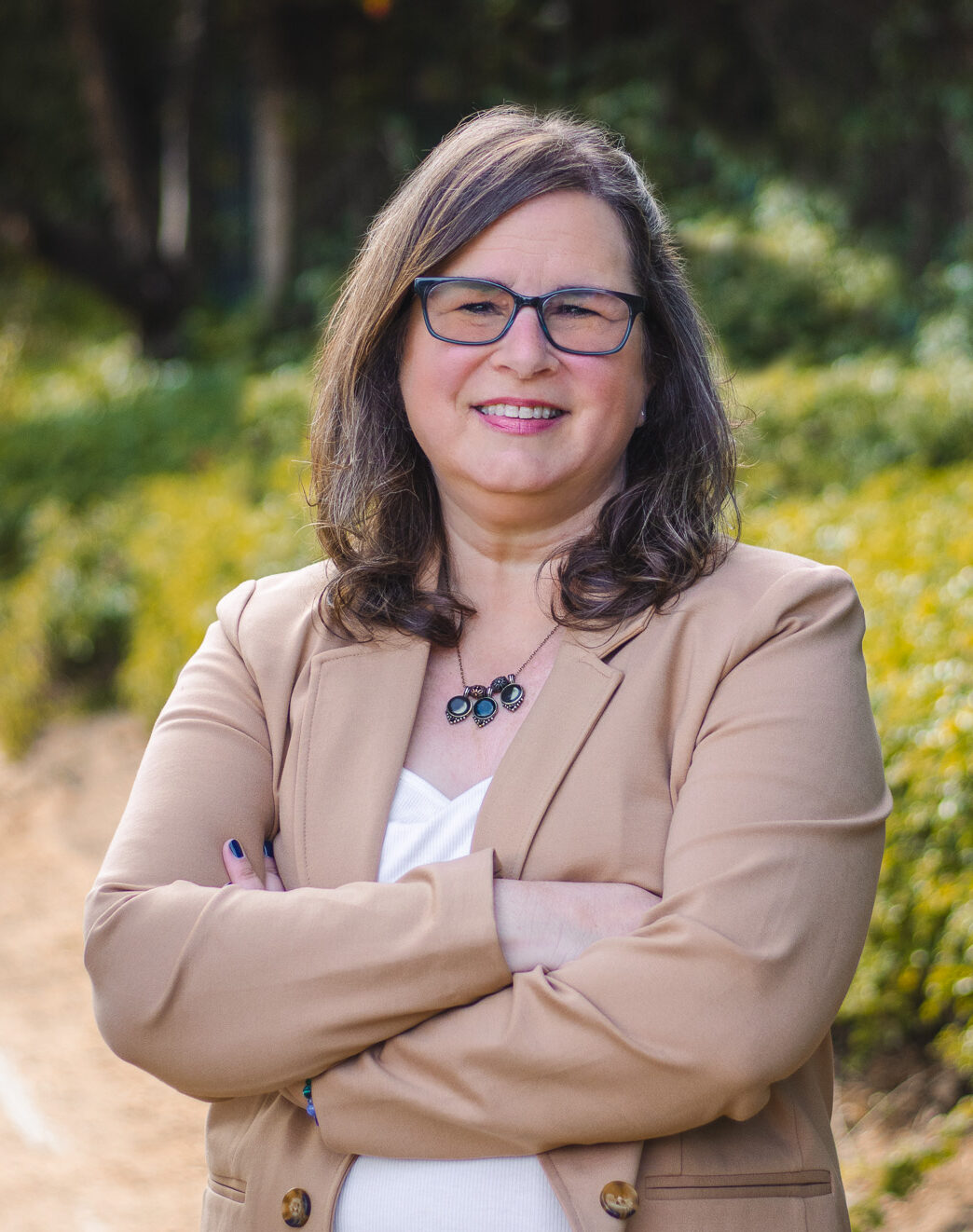Headshot of Mindy smiling with brown hair, dark rimmed glasses, and wearing a camel blazer and white shirt.