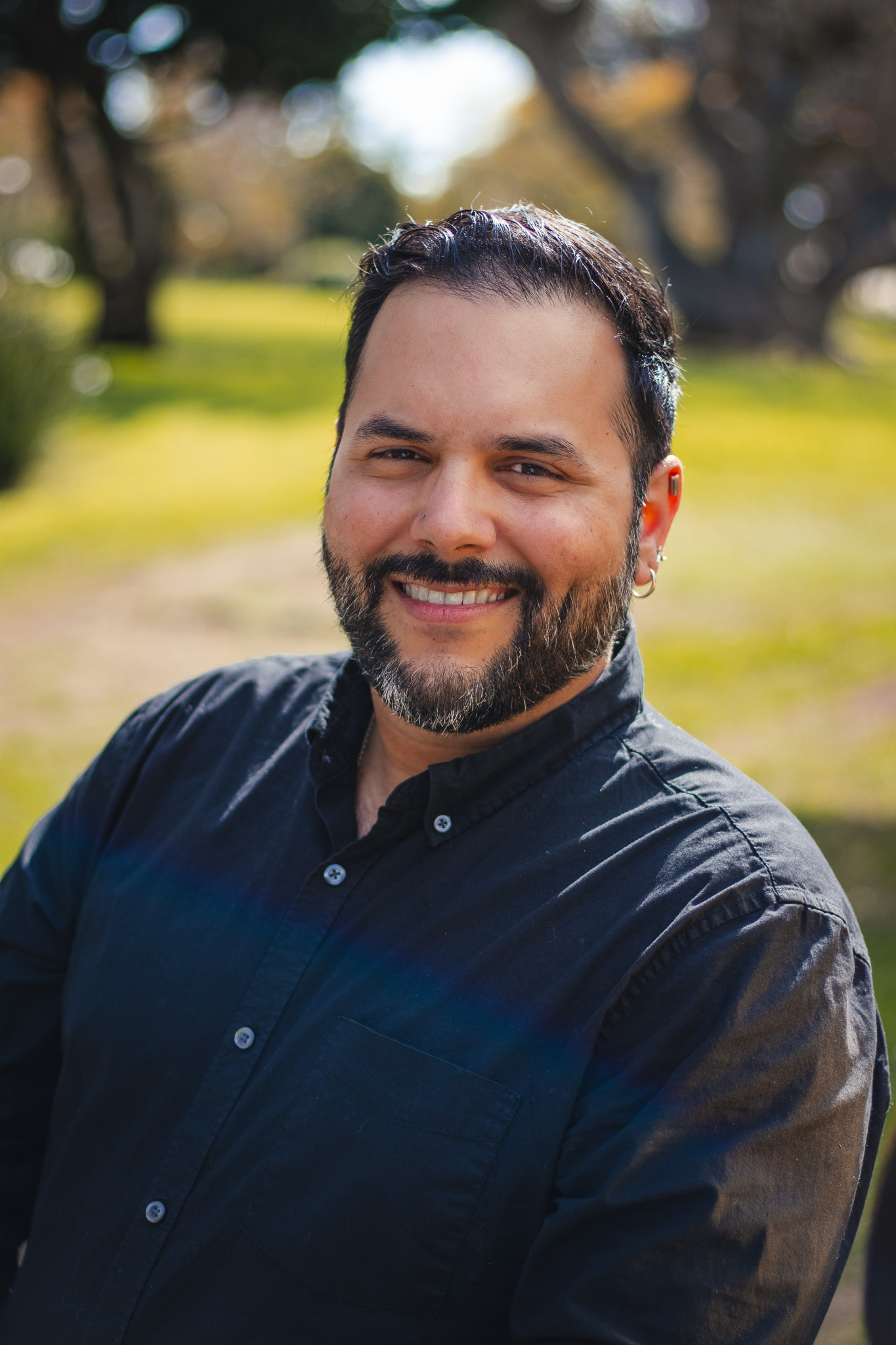 Headshot of Rolando smiling. Rolando has dark short hair, a neatly groomed beard, and is wearing a black button-up shirt.