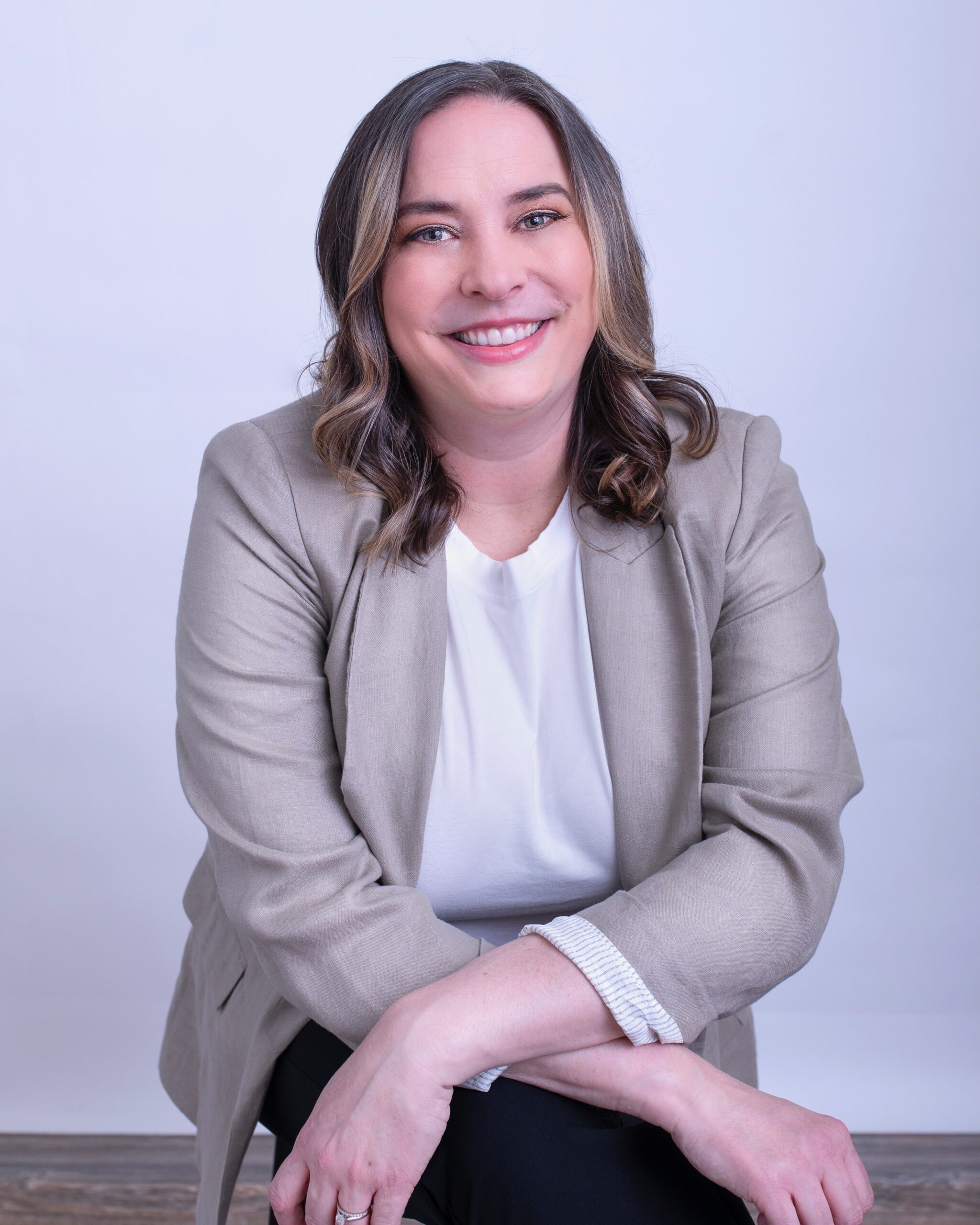 Kate Sonka has shoulder length brown hair, and grey blue eyes. She is smiling at the camera and wearing a tan blazer.