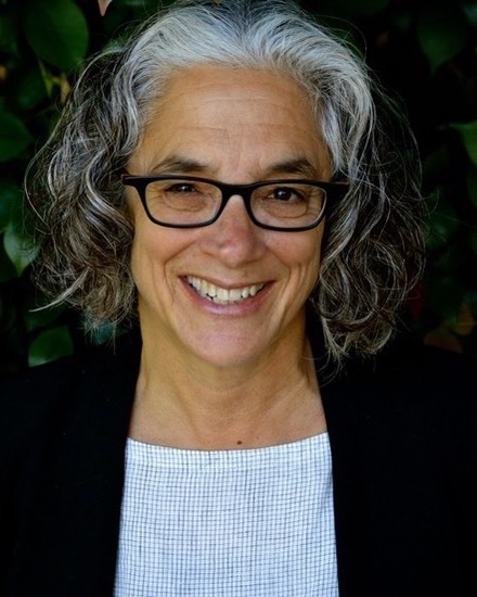 Headshot of Lainey Feingold. She has short gray curly hair and is wearing dark rimmed glasses. She is wearing a black blazer, white t shirt, and is smiling at the camera.