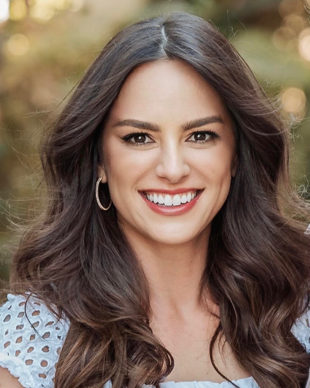 Liz has long dark brown hair, brown eyes, wearing a white top, and smiling at the camera.
