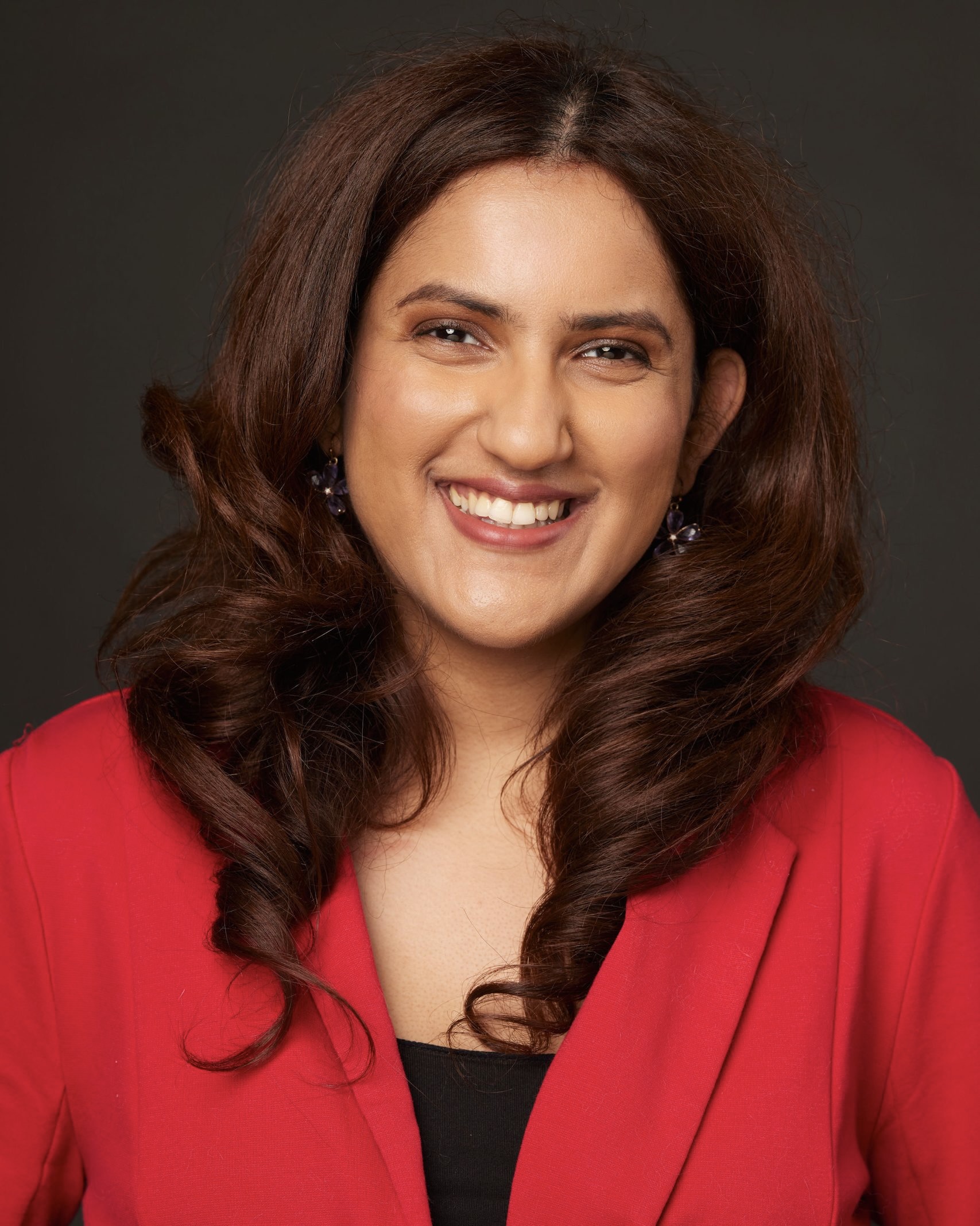 Meena has medium-toned skin and collarbone-length dark brown hair. She is smiling looking directly at the camera and wearing red blouse.