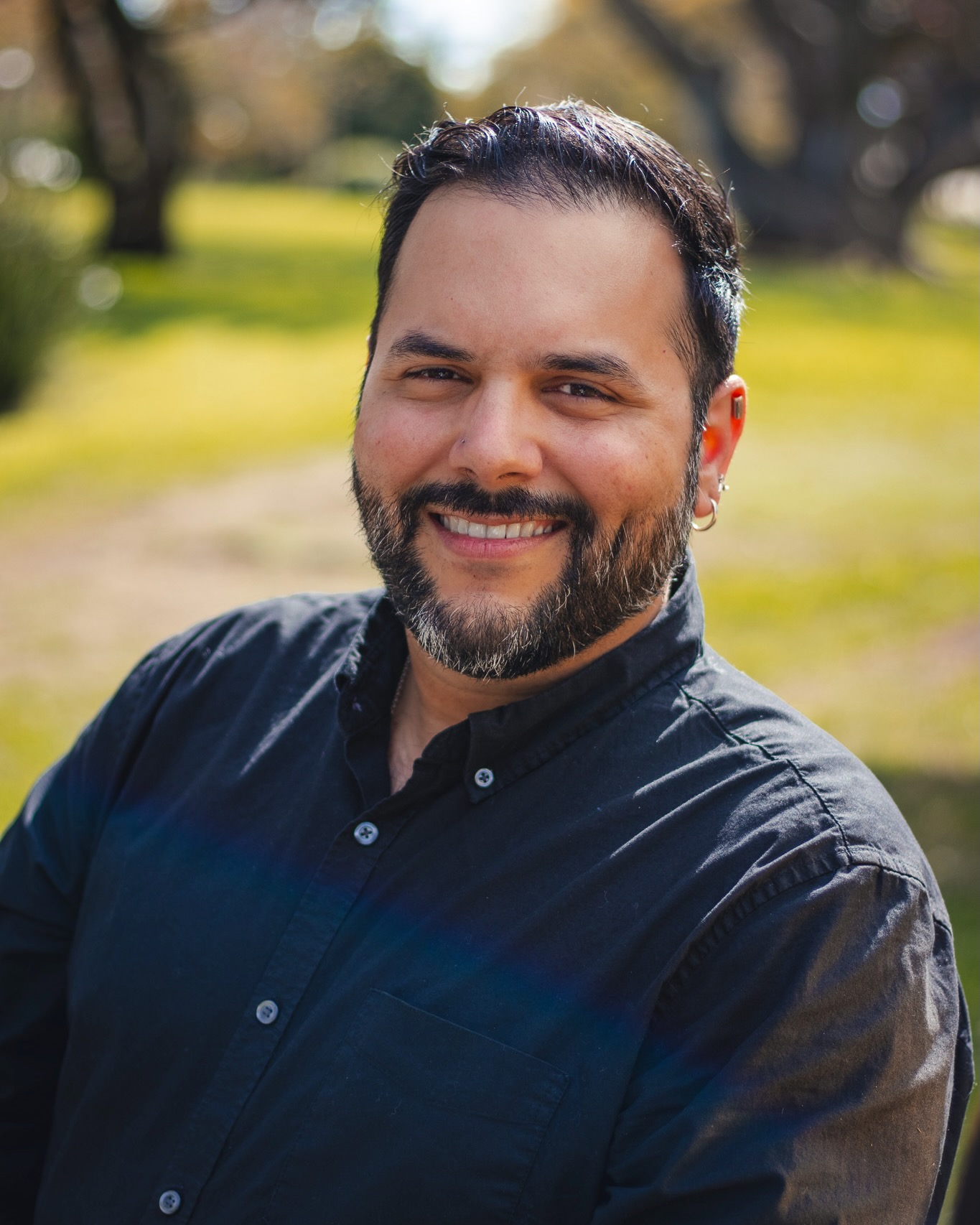 Headshot of Rolando smiling. Rolando has dark short hair, a neatly groomed beard, and is wearing a black button-up shirt.