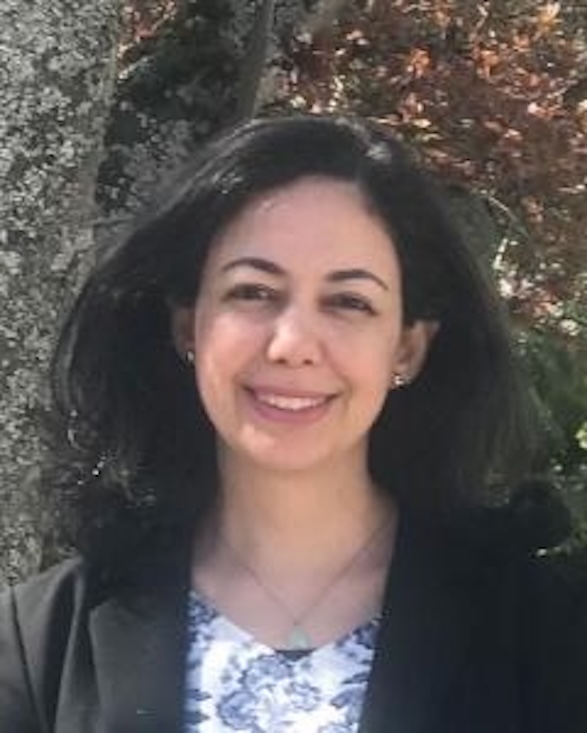 Yasmine has dark brown hair and brown eyes. She has a patterned top with a black cardigan on top. She is smiling at the camera
