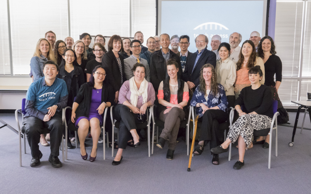 A large group of professionals pose for a photo in a conference room, with the Teach Access logo displayed on a presentation screen in the background.