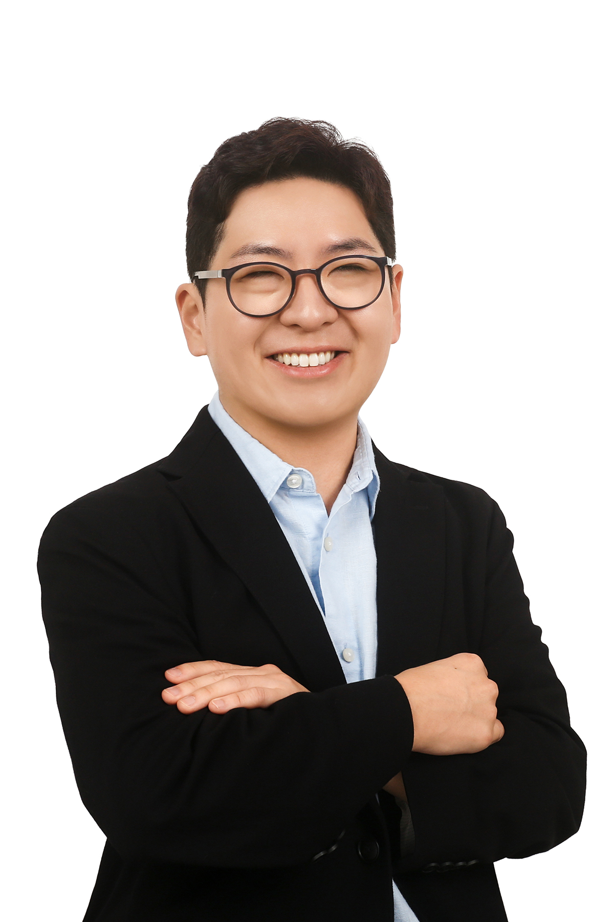 Professional headshot of Chris Yoon smiling, wearing round black glasses, a light blue button-down shirt, and a black blazer. They are standing with their arms crossed against a plain white background, looking directly at the camera with a friendly and confident expression.