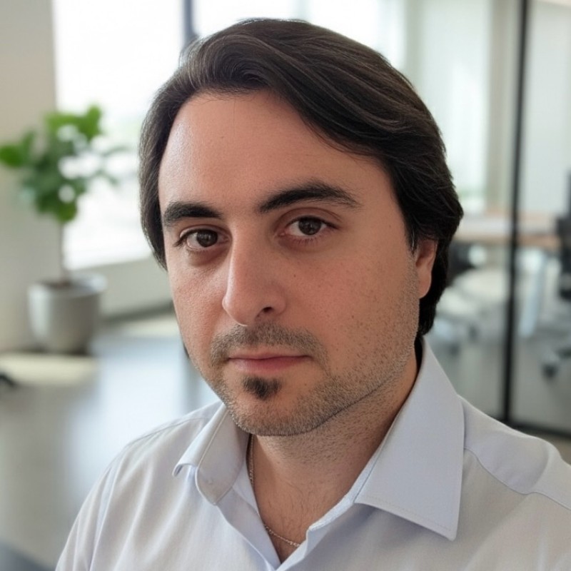 A portrait of Alihan Hadimlioglu, a man with dark hair and a goatee. He is wearing a light blue collared shirt and looking slightly off-camera, with a softly blurred office setting in the background.