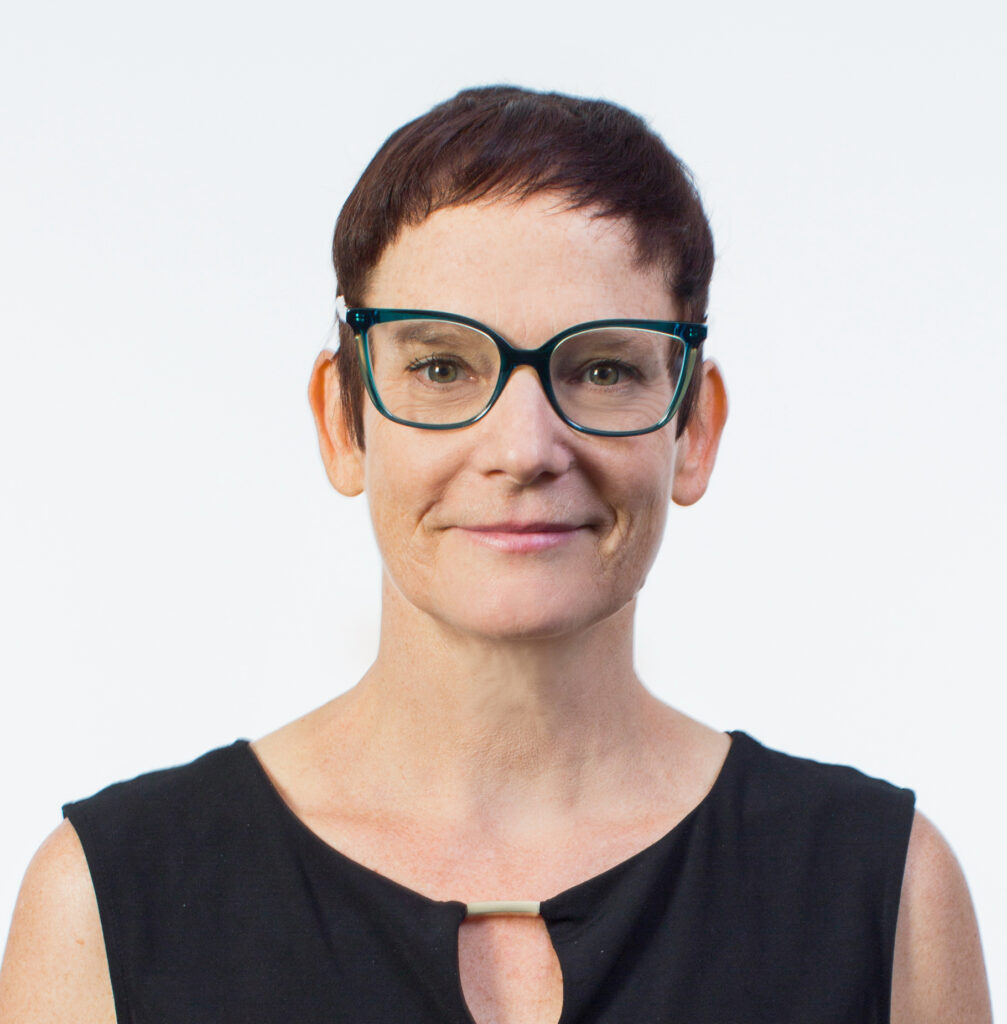 A headshot of Antonia Levy, a smiling woman with short reddish-brown hair. She is wearing green-rimmed glasses and a black sleeveless top with a small neckline cutout, set against a white background.