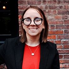 A portrait of Brianna Shuttleworth, a smiling woman with shoulder-length light brown hair. She is wearing dark round glasses, a ribbed red top, a simple necklace, and a black blazer, standing in front of a brick wall.