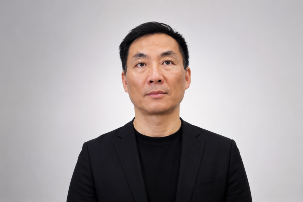 A professional headshot of Chen Wang, a man with short black hair and a neutral expression. He is wearing a black t-shirt underneath a black blazer, posed against a light grey background.