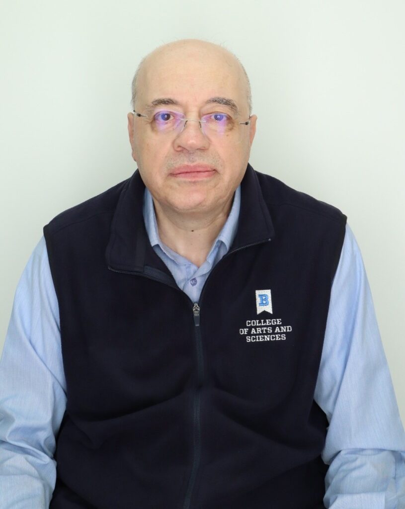 A headshot of Haitham Alkhateeb, a man with short, graying hair and wire-rimmed glasses. He is wearing a light blue button-down shirt under a dark blue fleece vest embroidered with the "College of Arts and Sciences" logo, against a white background.