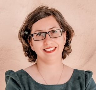 A portrait of Hajar Homayouni, a smiling woman with short brown hair. She is wearing rectangular black glasses, a delicate necklace, and a dark green top, set against a textured light beige/pink background.