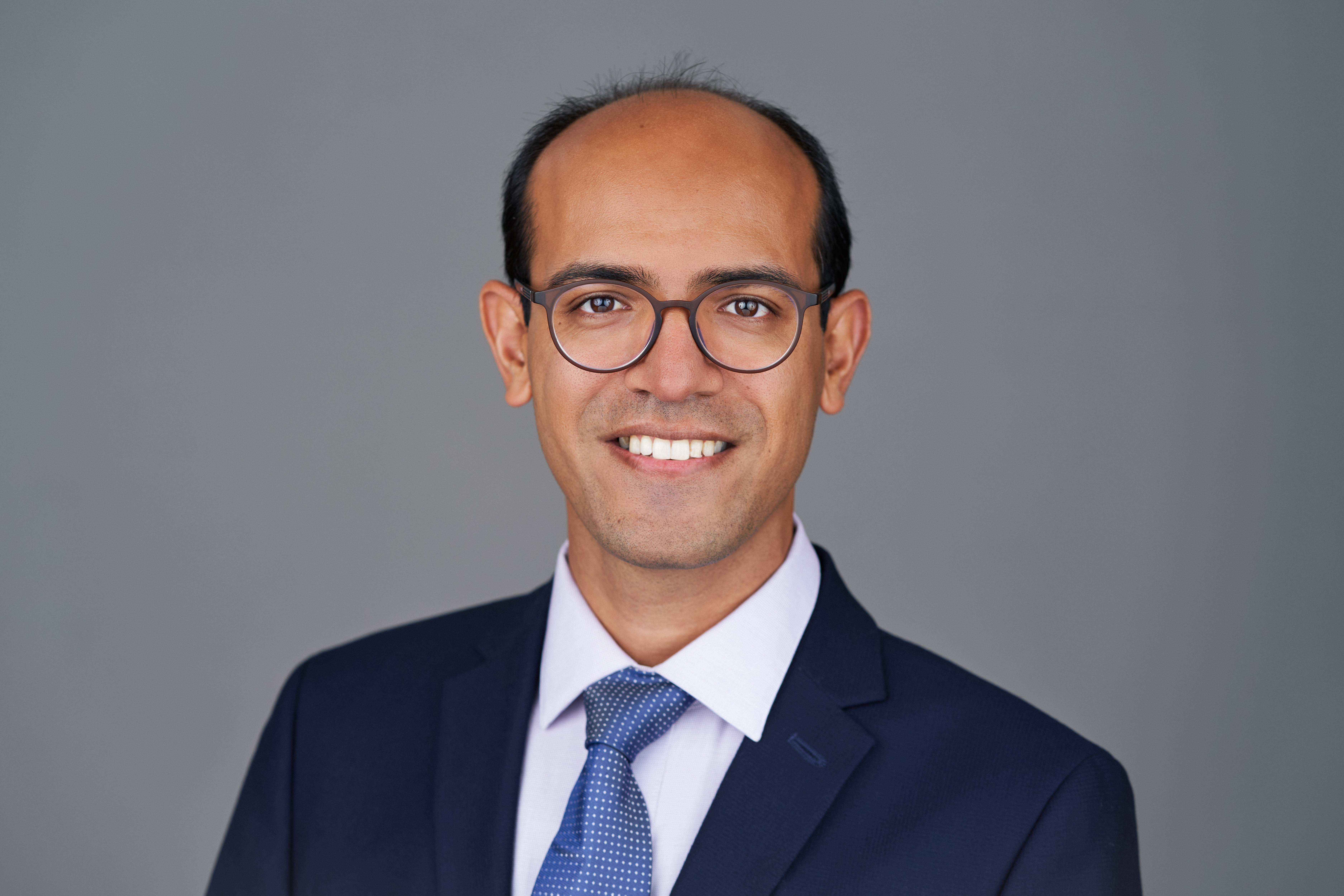 A professional headshot of Hossein Shirazi, a smiling man with a receding hairline and dark hair. He is wearing wire-rimmed round glasses, a light purple collared shirt, a dark blue suit jacket, and a patterned blue tie, posed against a grey background.