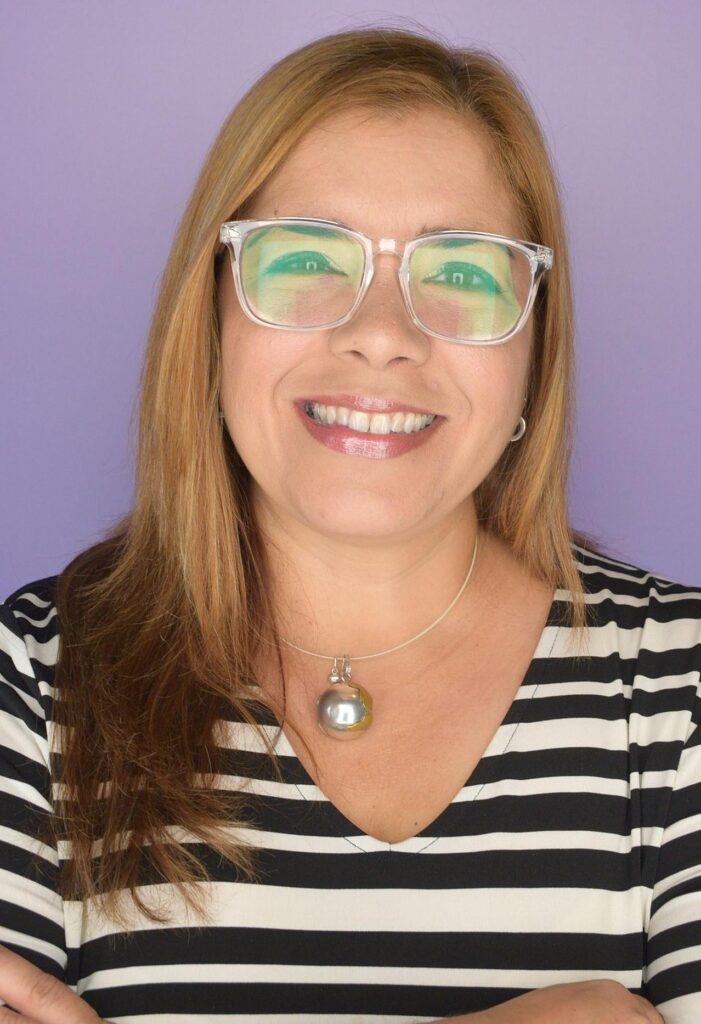 A headshot of Iliana Ballester Panelli, a smiling woman with shoulder-length light brown hair. She is wearing clear-rimmed glasses, a silver spherical pendant necklace, and a black and white striped top, against a solid light purple background.