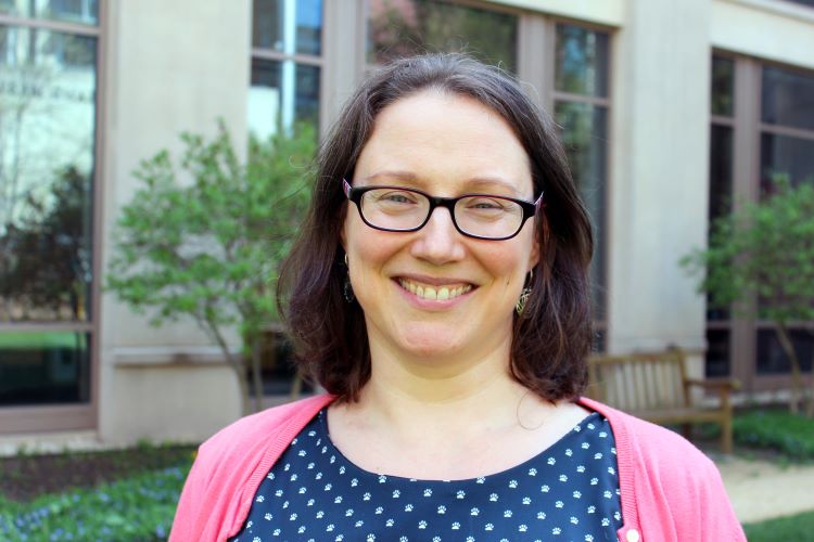 A portrait of Isabell May, a smiling woman with dark brown shoulder-length hair. She is wearing dark rectangular glasses, a dark blue shirt with small white paw prints, and a bright pink cardigan, standing outdoors in front of a building with large windows.