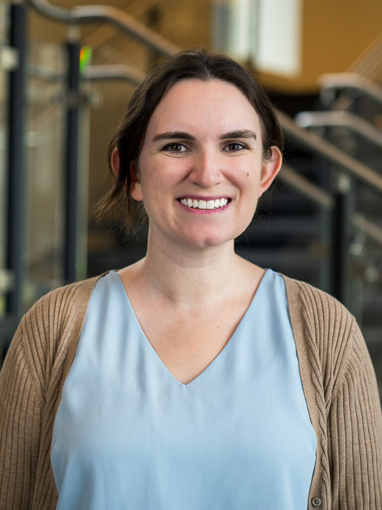 A headshot of Jennifer Kowalski, a smiling woman with dark brown hair pulled back. She is wearing a light blue v-neck top under a tan ribbed cardigan, posed indoors with a softly blurred staircase in the background.
