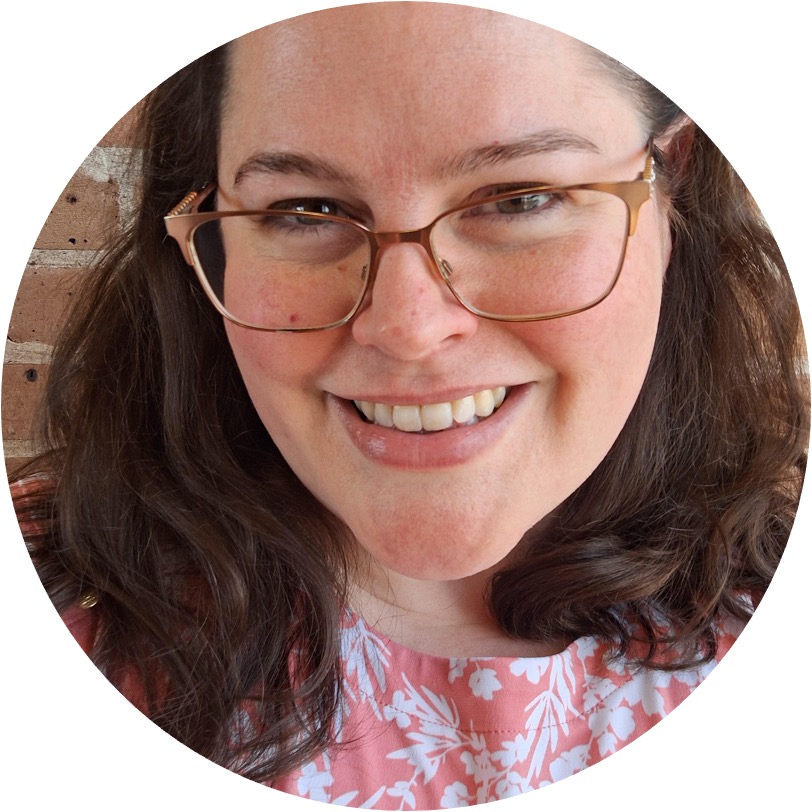 A close-up, circular-cropped portrait of Jessica Frances Handley, a smiling woman with wavy brown hair. She is wearing bronze-rimmed glasses and a pink top with white floral patterns, set against a blurred brick wall.