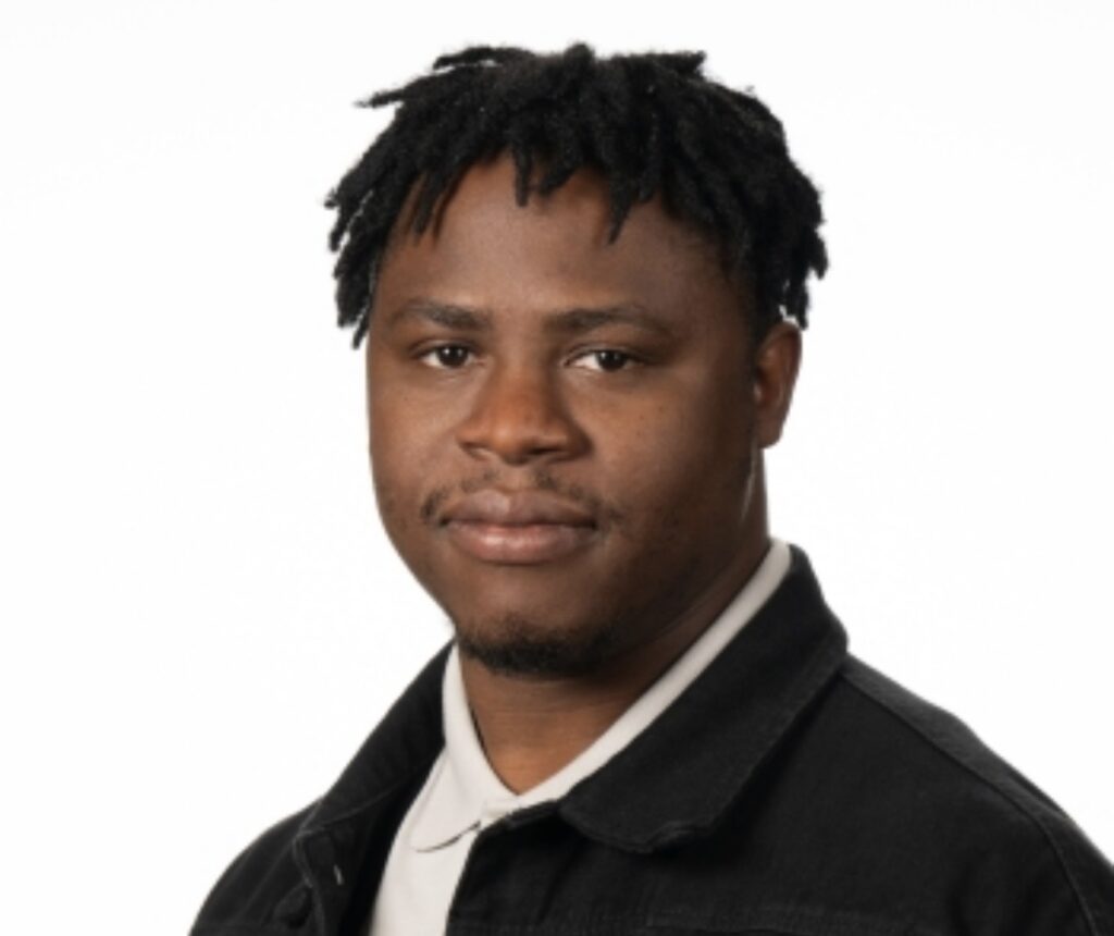 A professional headshot of Jude Okolie, a man with short, dark locs. He has a slight smile and is wearing a black jacket over a white collared shirt, posed against a solid white background.