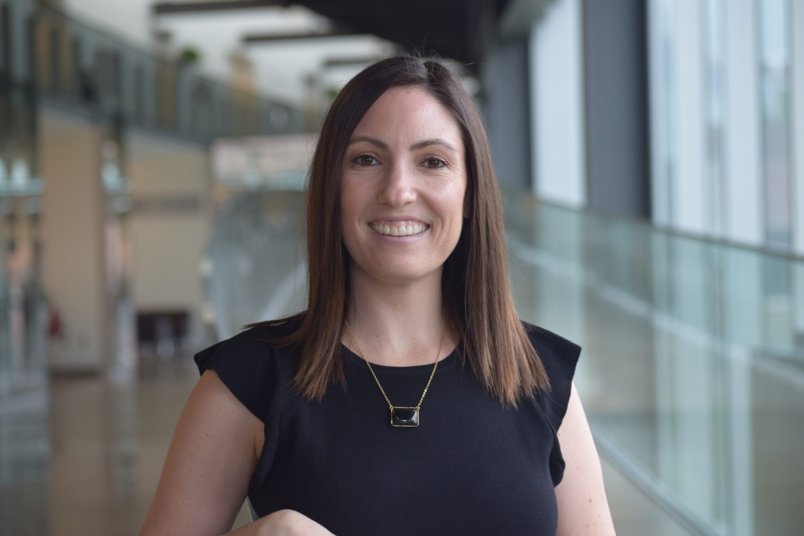 A portrait of Kristen Cole, a smiling woman with straight brown hair. She is wearing a black short-sleeved top and a rectangular dark pendant necklace with gold trim, standing in a brightly lit, modern indoor setting with glass walkways behind her.