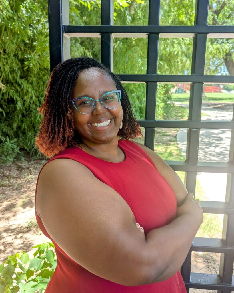 A portrait of Leila Lester, a smiling woman with dark locs featuring reddish tips. She is wearing blue-rimmed cat-eye glasses and a sleeveless red dress, standing outdoors with her arms crossed next to a black metal trellis, with green trees and a park area in the background.