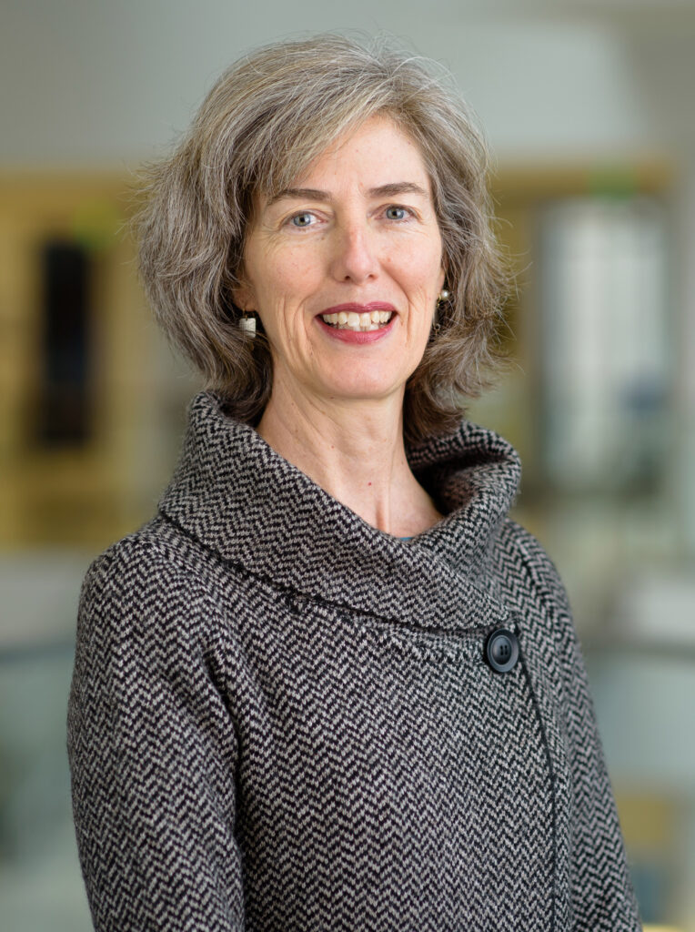 A portrait of Marie Frank, a smiling woman with short, wavy gray hair. She is wearing a textured, high-collared, black-and-white herringbone pattern coat with a large black button, standing in a softly blurred indoor space.
