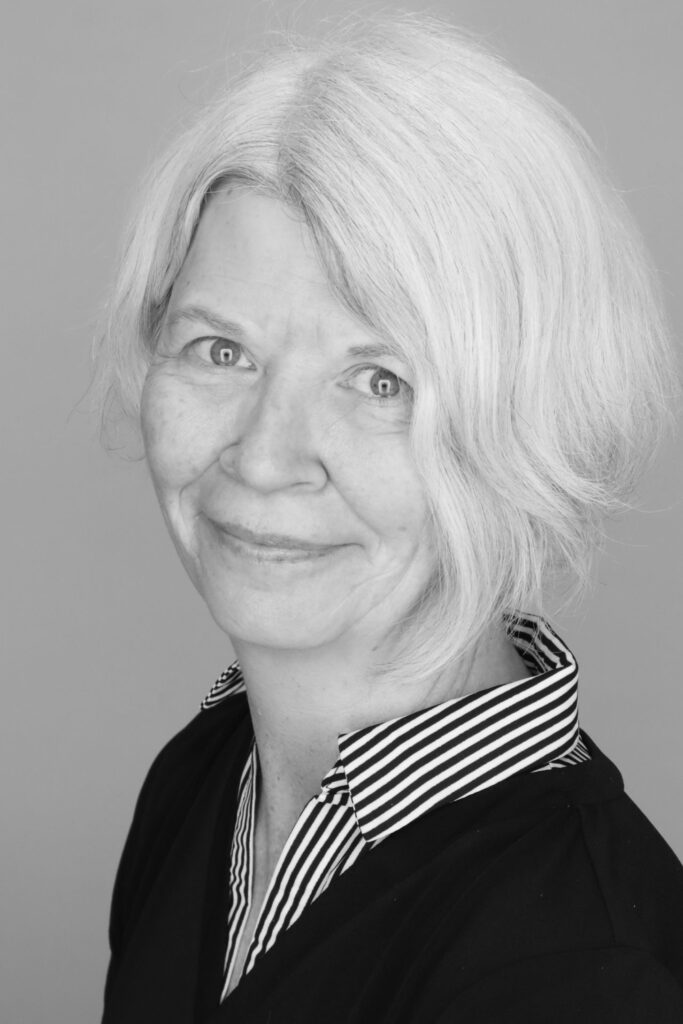 A black-and-white headshot of Mary-Colleen Jenkins, a smiling woman with short, light-colored hair. She is wearing a dark top layered over a black-and-white striped collared shirt, posed against a solid grey background.