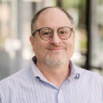A portrait of Michael Canale, a smiling man with a receding hairline and a gray goatee. He is wearing round brown glasses and a light blue striped button-down shirt, set against a softly blurred background.
