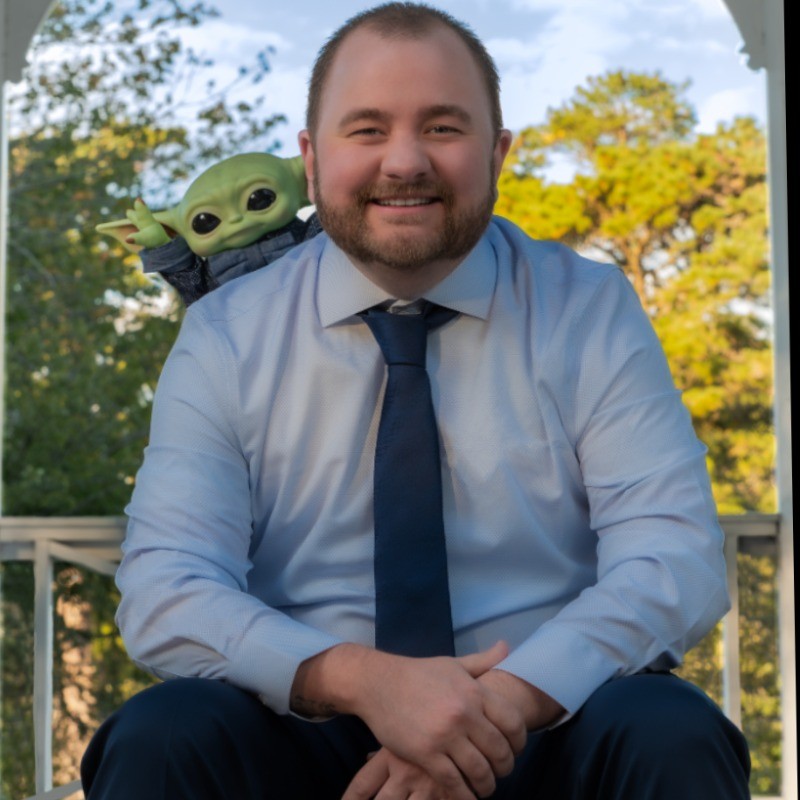 A portrait of Nicholas Alexander, a smiling man with short brown hair and a beard. He is wearing a light blue dress shirt and a dark blue tie. A small Baby Yoda (Grogu) plush toy is sitting on his right shoulder. He is posed outdoors with a white porch railing and green trees in the background.