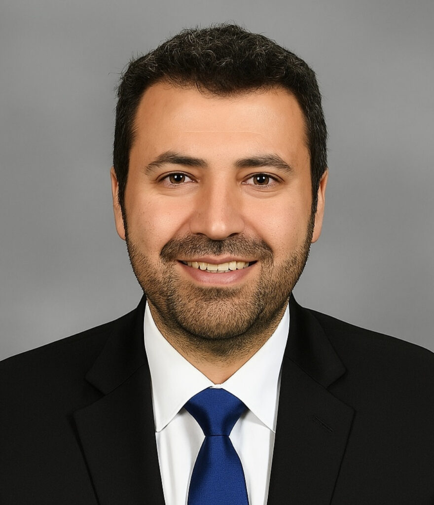 A professional headshot of Nuri Yilmazer, a smiling man with short dark hair and a beard. He is wearing a black suit jacket, a white collared shirt, and a solid blue tie, posed against a solid gray background.