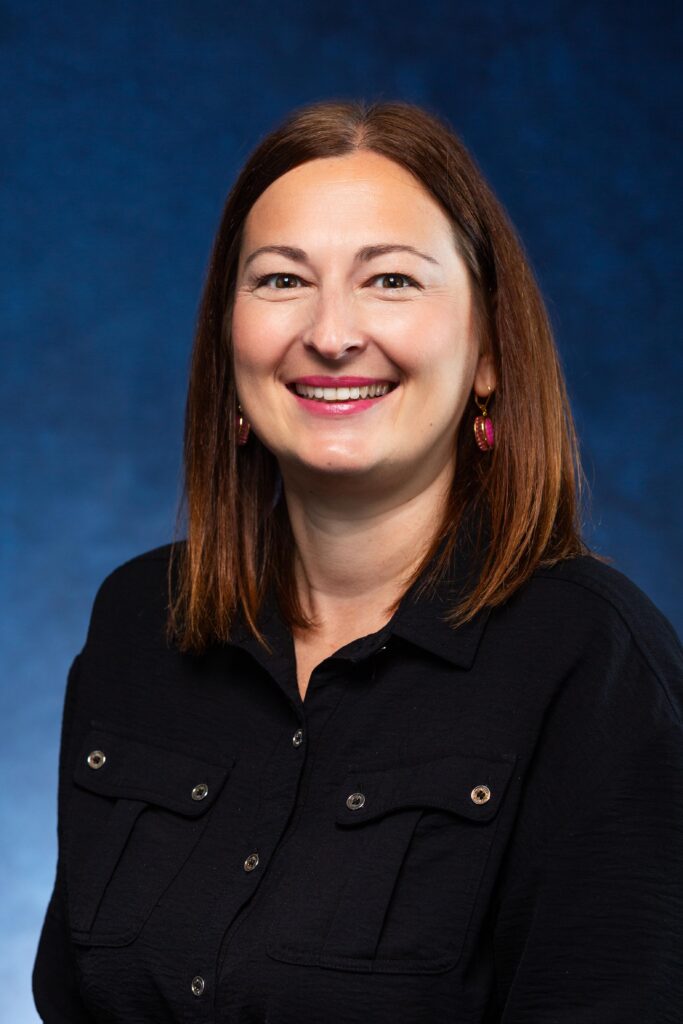 A headshot of Renata Endres, a smiling woman with straight, shoulder-length brown hair. She is wearing small earrings and a black button-down shirt with chest pockets, set against a mottled blue background.