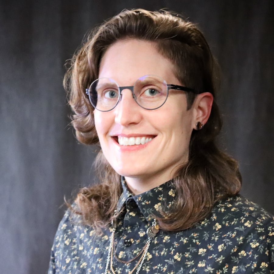 A headshot of Rua Mae Williams, a smiling person with wavy brown hair that is shorter on one side. They are wearing round wire-rimmed glasses, a dark floral print button-up shirt, and layered necklaces, set against a dark gray background.