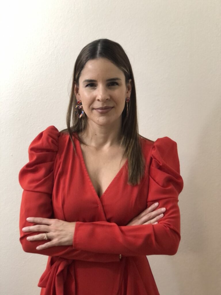 A portrait of Ruth Mercado Cruz, a woman with long straight brown hair, standing with her arms crossed. She is wearing a bright red long-sleeved dress with gathered shoulders and colorful dangling earrings, set against a plain white wall.