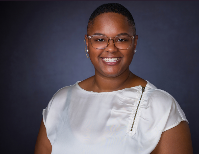A portrait of Shavonda Jackson, a smiling woman with very short hair. She is wearing clear-rimmed glasses, a white short-sleeved top with a zipper detail on the left shoulder, and pearl stud earrings, set against a dark gray background.