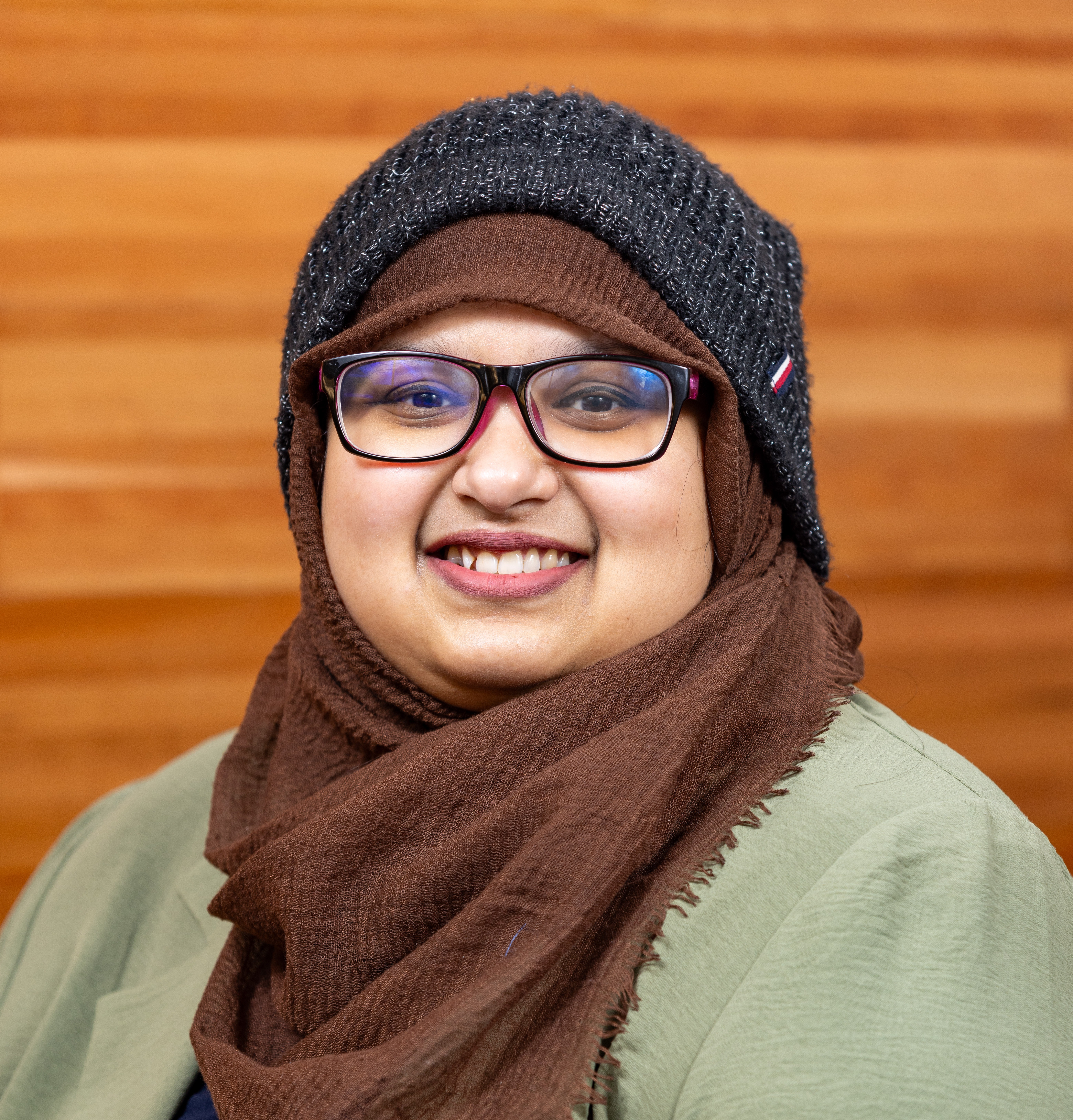 A portrait of Tamanna Motahar, a smiling woman wearing a textured brown hijab and a dark knit winter beanie. She is wearing dark-rimmed glasses and a light olive green jacket, posed in front of a warm, wooden paneled wall.