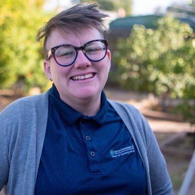 A portrait of Tracy Stuntz, a smiling person with short brown hair. They are wearing round tortoiseshell glasses, a dark blue polo shirt with a logo, and a light gray cardigan, standing outdoors with a softly blurred background of green foliage.