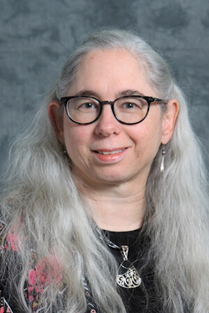 A professional headshot of Suzanne David, smiling with long, wavy gray hair. They are wearing dark round-rimmed glasses, a black top, a patterned outer layer, and a silver pendant necklace, set against a mottled gray background.