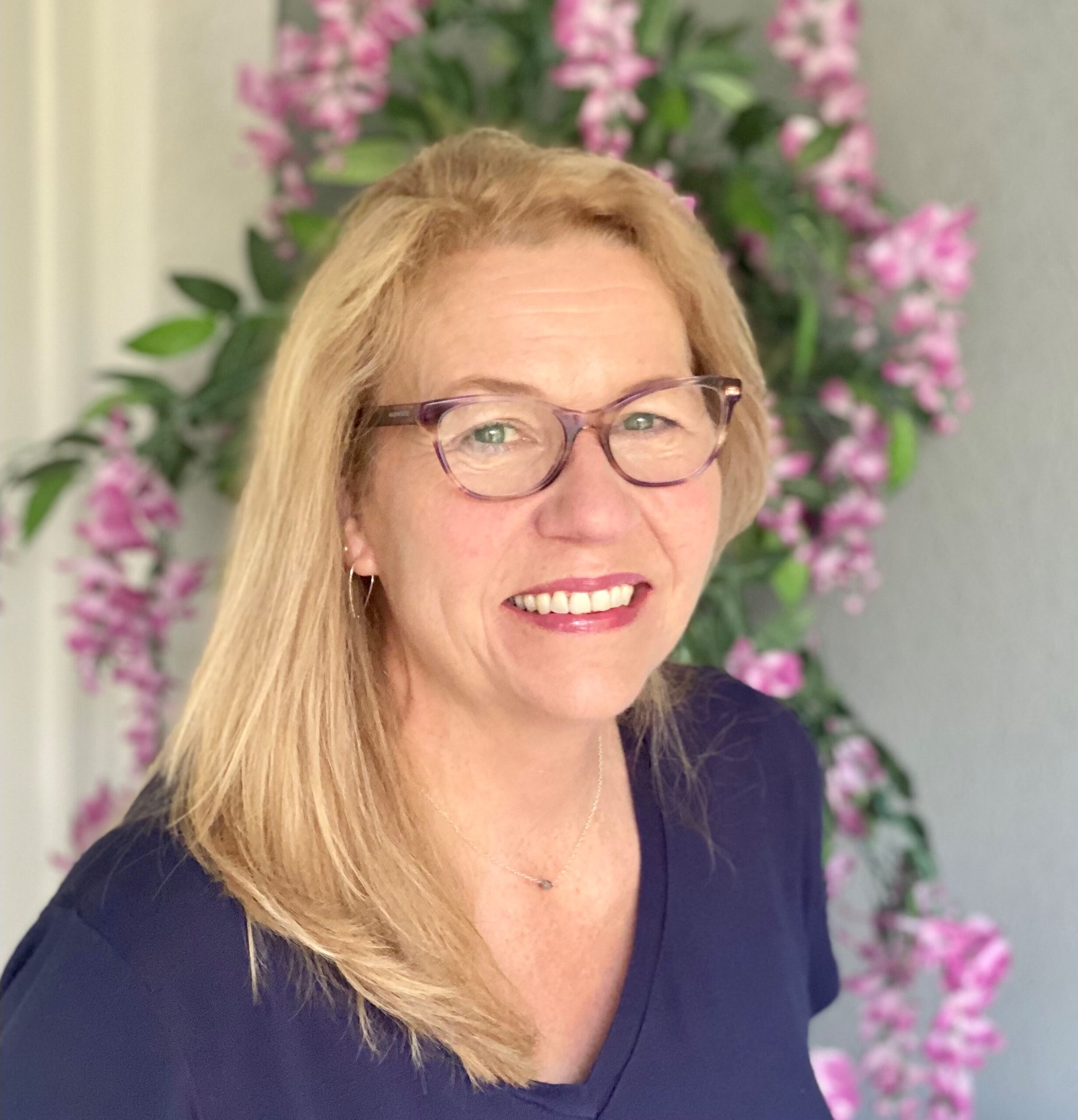 A photo of Lori Samuels smiling. She is a woman with blond hair and purple-framed glasses stands against a backdrop of pink flowers, wearing a dark shirt.