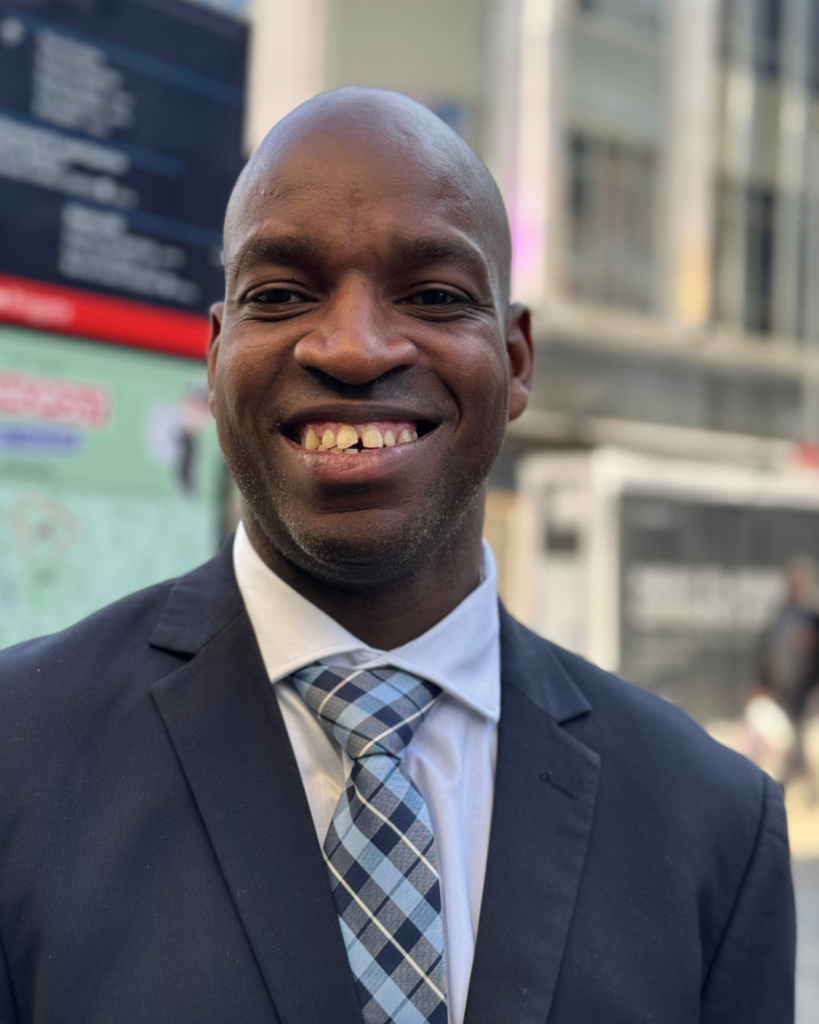 A close-up portrait of Edmore Masendeke, a smiling bald Black man wearing a dark suit jacket, white dress shirt, and blue-gray plaid tie. He faces the camera and fills most of the frame, with a blurred urban streetscape and a partially visible colorful sign in the background.